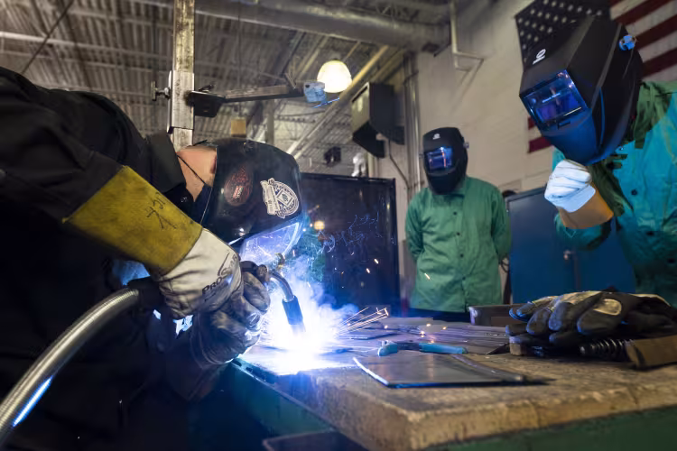 Three students wearing welding masks and jackets work together on a project.