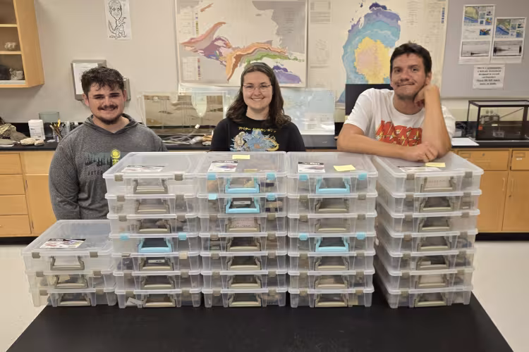 Three students stand behind stacks of plastic containers containing fossils.