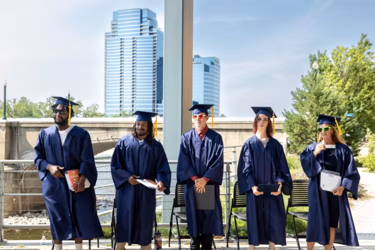 Five landscape apprentices in their caps and gowns stand with the iconic Grand Rapids skyline in the background.