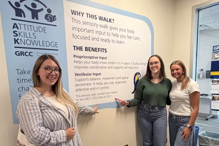 Three students stand outside the OTA department showing off a sign on the wall that explains the sensory walk.