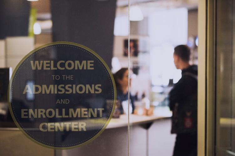 The glass wall of the Enrollment Center with a sign on the front and students in the background.