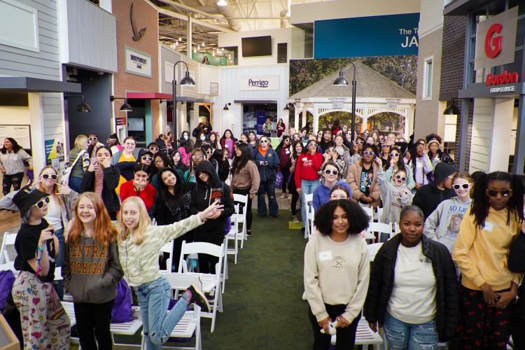 Dozens of children pose, wearing sunglasses, at an indoor display area featuring local businesses.