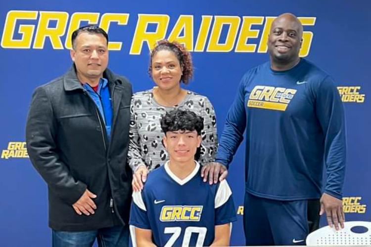 GRCC Jaret Jaramillo sits at a table in front of three people, all are standing in front of a GRCC Raiders banner.