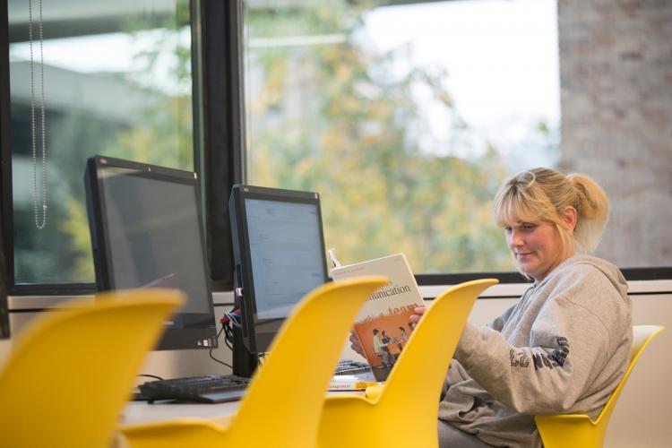 A female GRCC student sitting in a computer lab, looking at a text book.