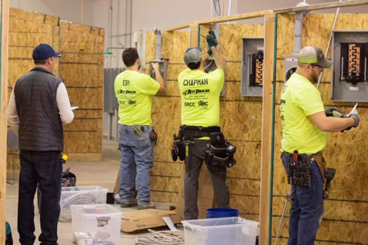 A judge, holding a clipboard, watches students perform an electrical installation at Tassell M-TEC.
