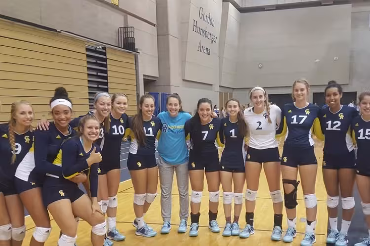 Volleyball team members stand in a row in the fieldhouse with their arms around each other.