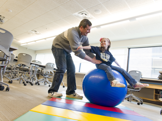 An Occupational Therapy Assistant helps a child find balance on a yoga ball.