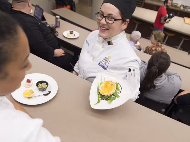 A student chef presenting a dish to a student.