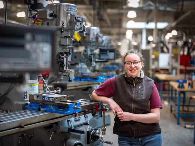 A person stands in a shop and leans against heavy machinery. 