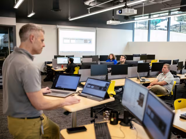 An instructor stands near four computer monitors in a GRCC class. 