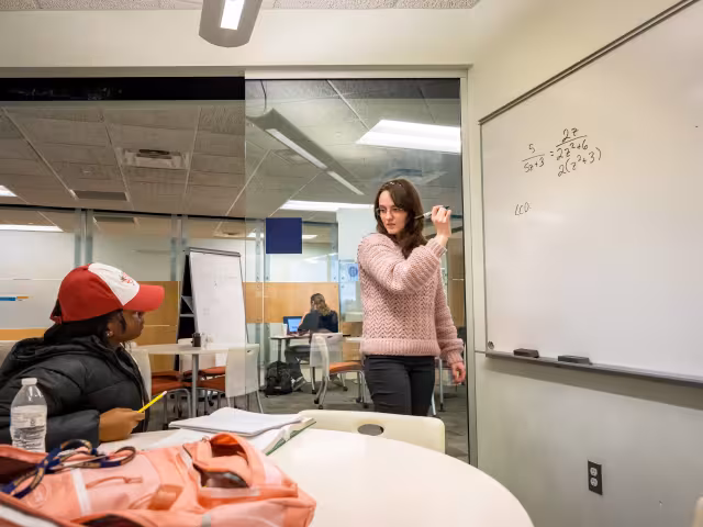 A math tutor writes out a problem on a white board in a study room.
