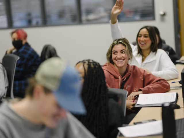 A student in a math class raises her hand while the students around her are smiling.