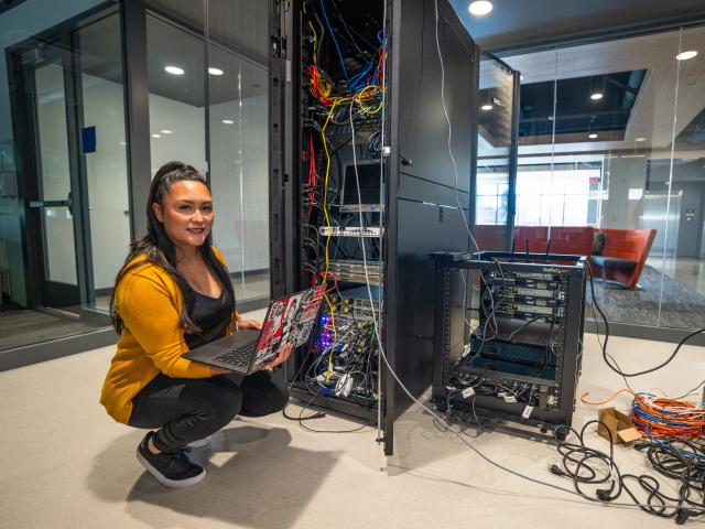 A student holds a laptop in front of a server rack full of wires.