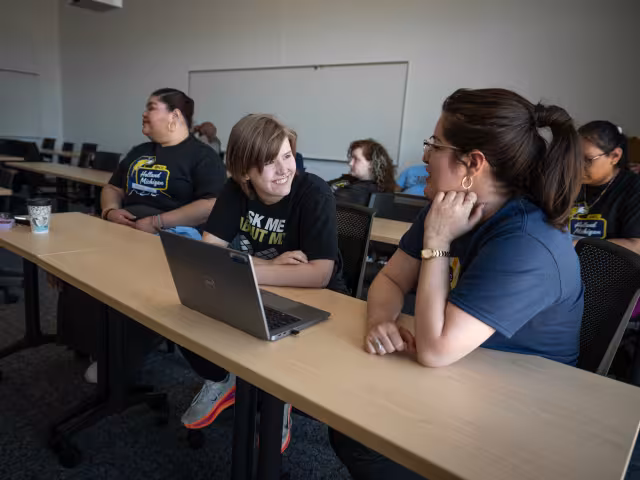 Two students sit at a laptop, talking. 